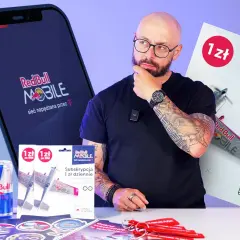 Mężczyzna z tatuażami zastanawia się nad ofertą Red Bull MOBILE. Na stole leżą puszki Red Bull, karty SIM i gadżety.