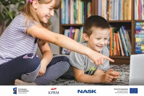 Dzieci z uśmiechem odkrywają świat gier cyfrowych na laptopie, siedząc na dywanie przed regałem pełnym książek.