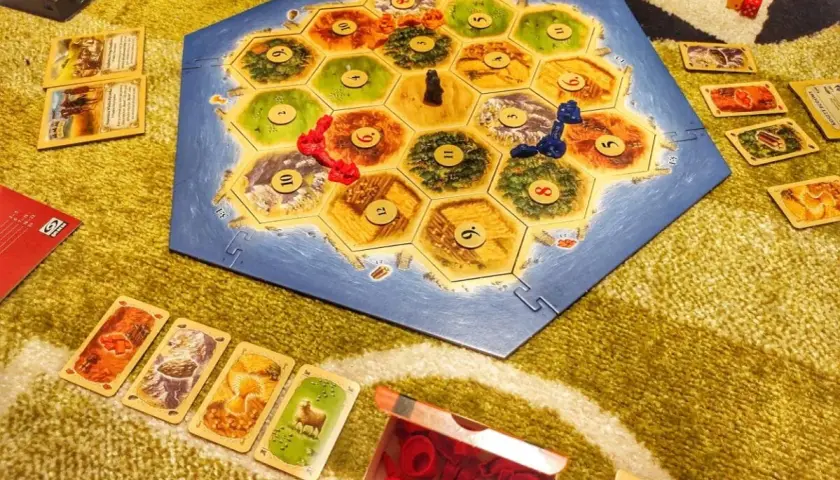 Catan wersja podróżna na dywanie. Plansza z wyspą, karty surowców i pionki.